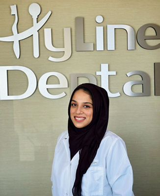 The image shows a person standing in front of a sign that reads  Hyloline Dental.  The individual is smiling and appears to be wearing a white lab coat, suggesting they may work in the dental field.