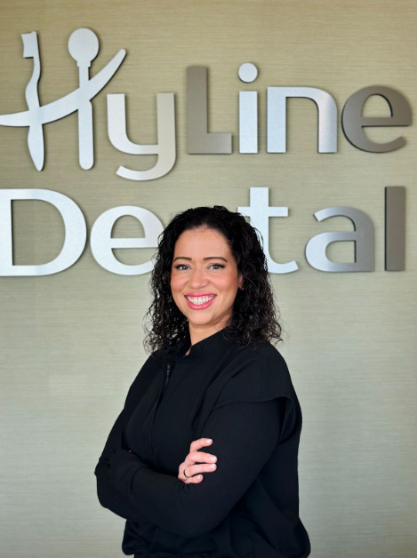 The image is a photograph of a woman standing in front of an office sign that reads  Hylleline Dental.  She appears to be posing for the photo, with her hands on her hips and a slight smile.