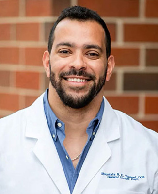 A smiling man in a white lab coat stands against a brick wall, posing for the camera.