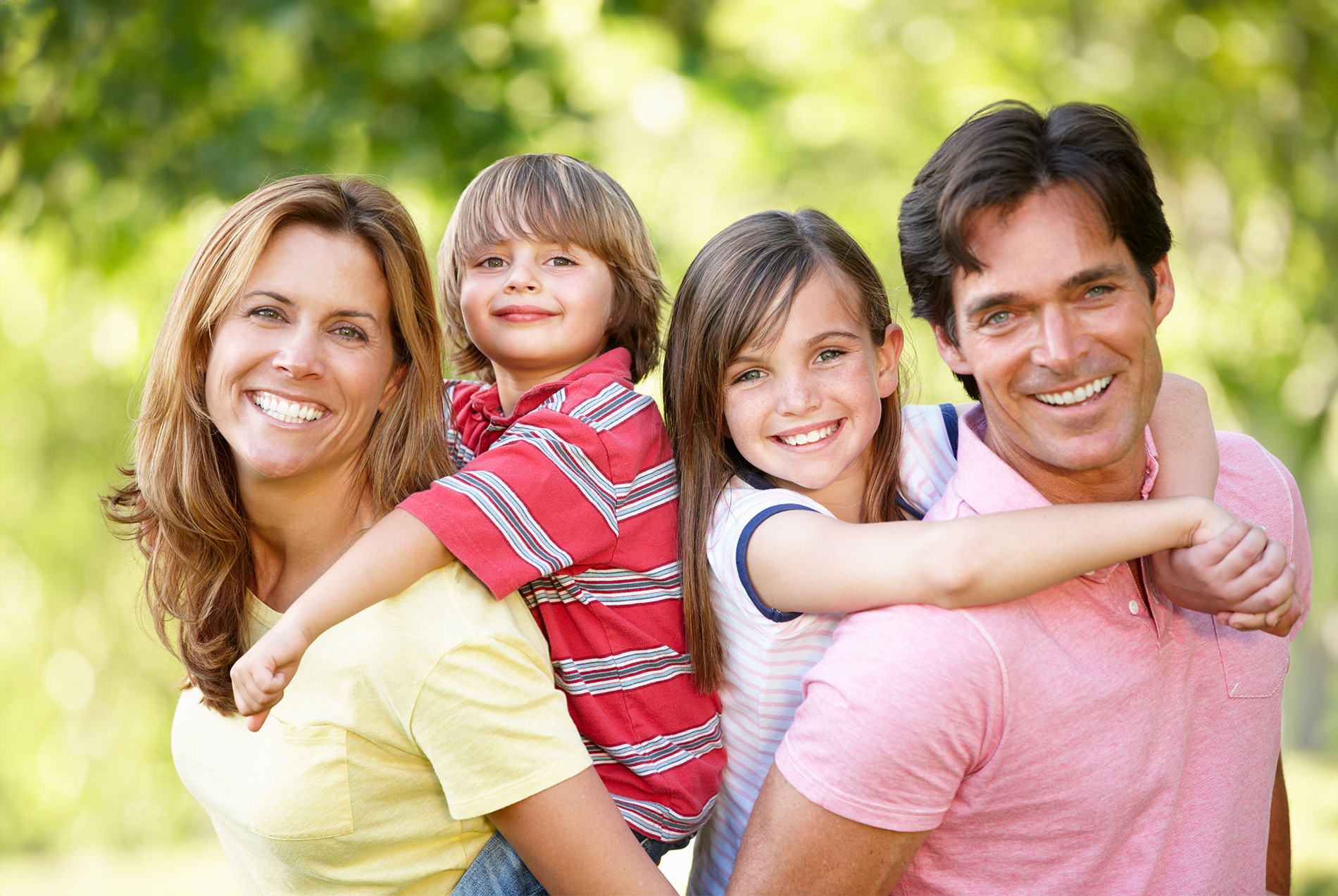 The image is a family portrait featuring four people  an adult couple and their two children, posing together outdoors with trees in the background.