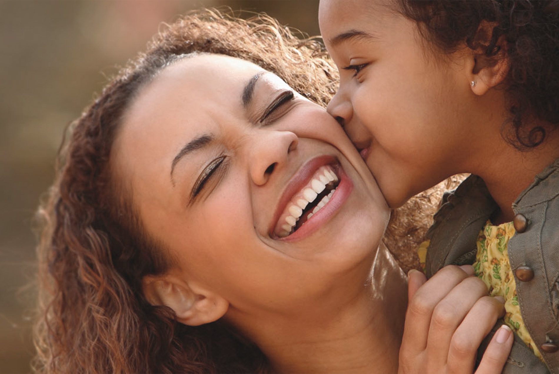 The image shows a woman with her mouth open, laughing heartily while hugging a child who is also smiling. They are both outdoors, and the woman appears to be in mid-motion, suggesting a joyful moment captured between them.