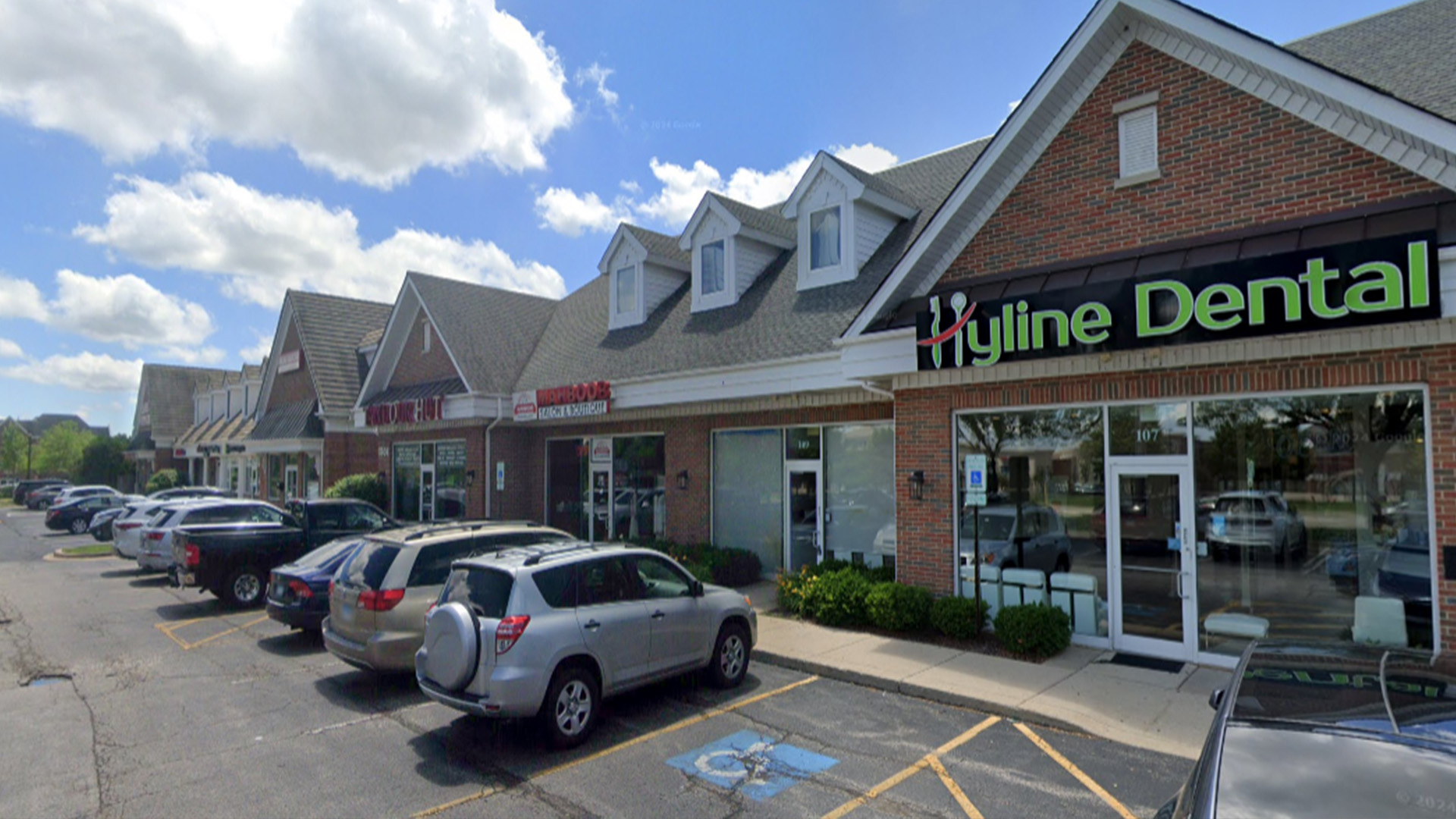 The image is a photograph of an outdoor scene featuring a two-story building with the sign  Hygiene Dental  on its facade, adjacent to a parking lot with several parked cars.