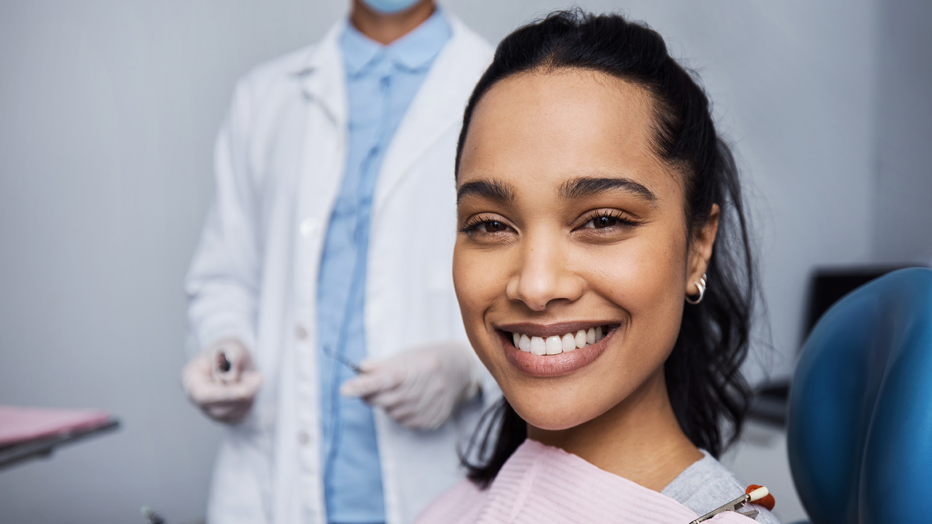 In the image, a woman is seated in a dental chair with a smile on her face. She is wearing a blue surgical mask and has white teeth. A dentist stands behind her, also wearing a blue mask and holding a dental tool. They both appear to be engaged in a dental examination or procedure.