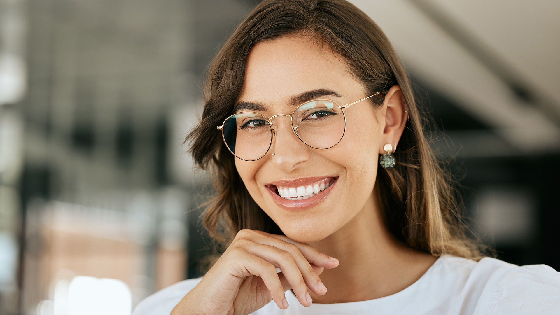 The image is a portrait of a smiling woman with glasses, wearing a white top, and her hand resting on her chin.