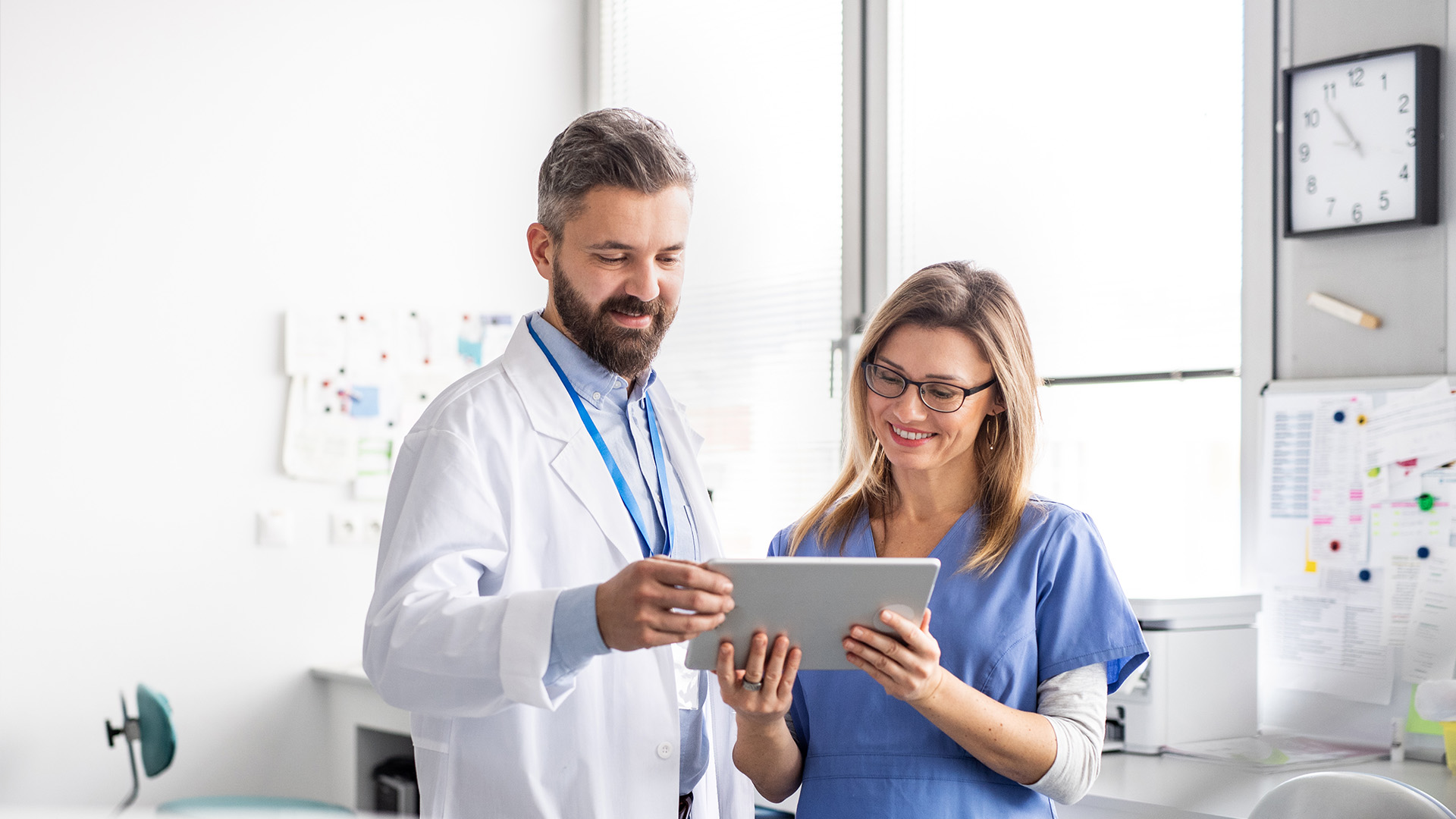 A man in a white lab coat and a woman in scrubs, both holding tablets, are standing in an office setting with a window behind them.