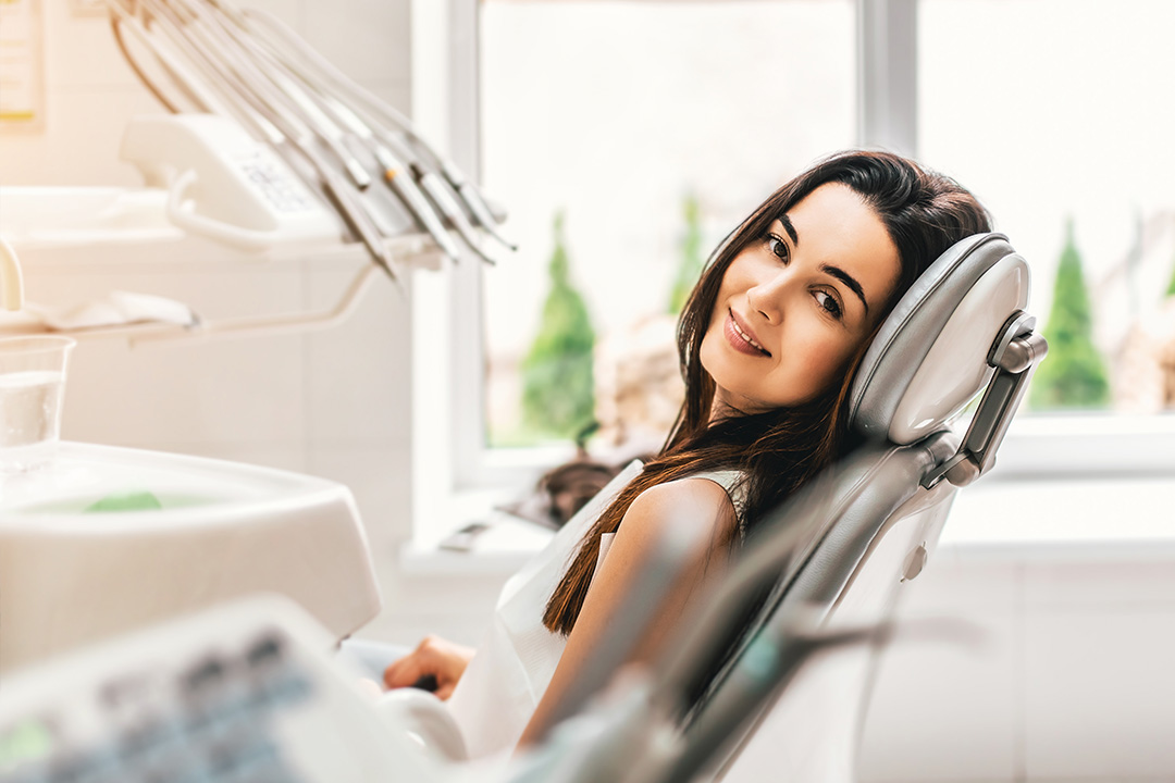 A woman sitting in a dental chair, smiling at the camera, with a dental office setting visible behind her.