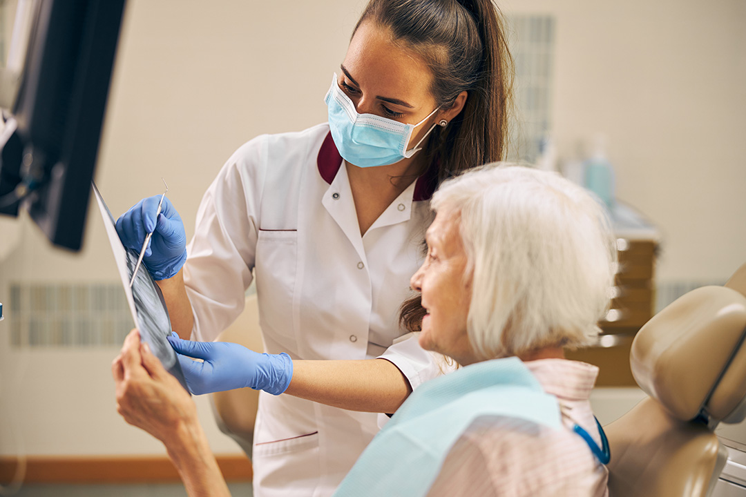 A dentist in a white lab coat assisting an elderly woman with a dental appointment, both wearing face masks.