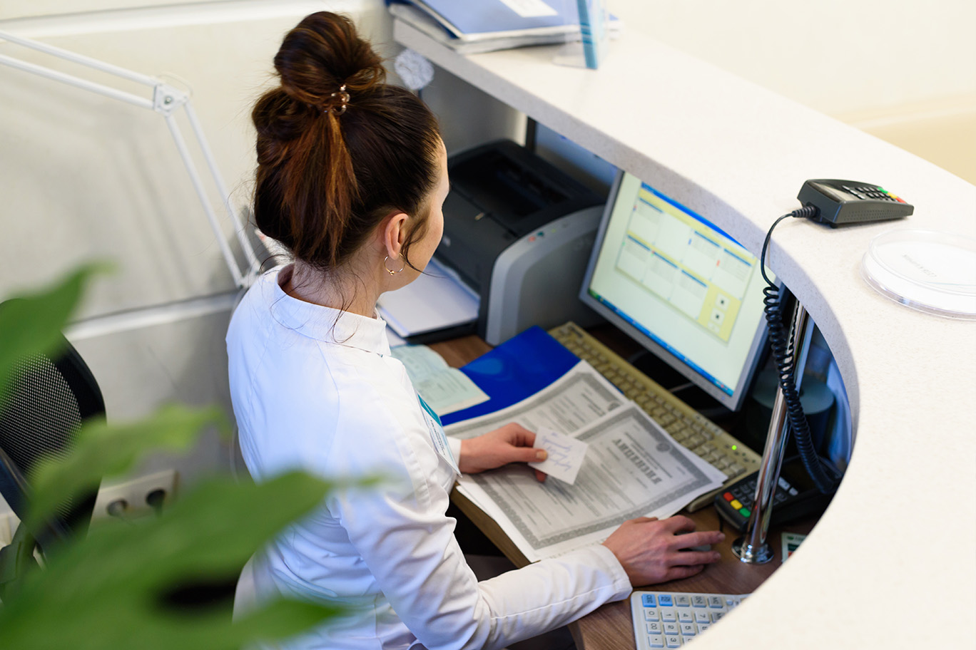 A woman in a white shirt and black pants is standing at a counter with a computer monitor, keyboard, and mouse.