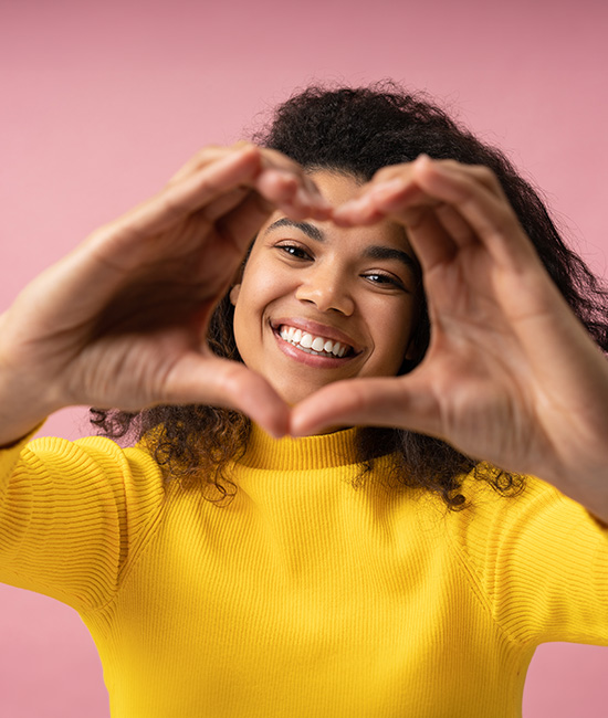 The image shows a person making a heart shape with their hands, smiling and looking directly at the camera against a pink background.
