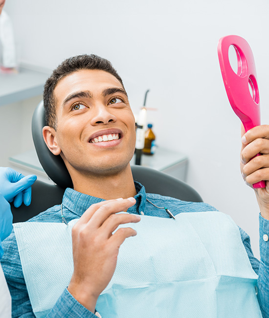 The image shows a man sitting in a dental chair, smiling broadly, holding a pink toothbrush in his hand, with a dental hygienist standing to the right, wearing gloves and holding a dental mirror.