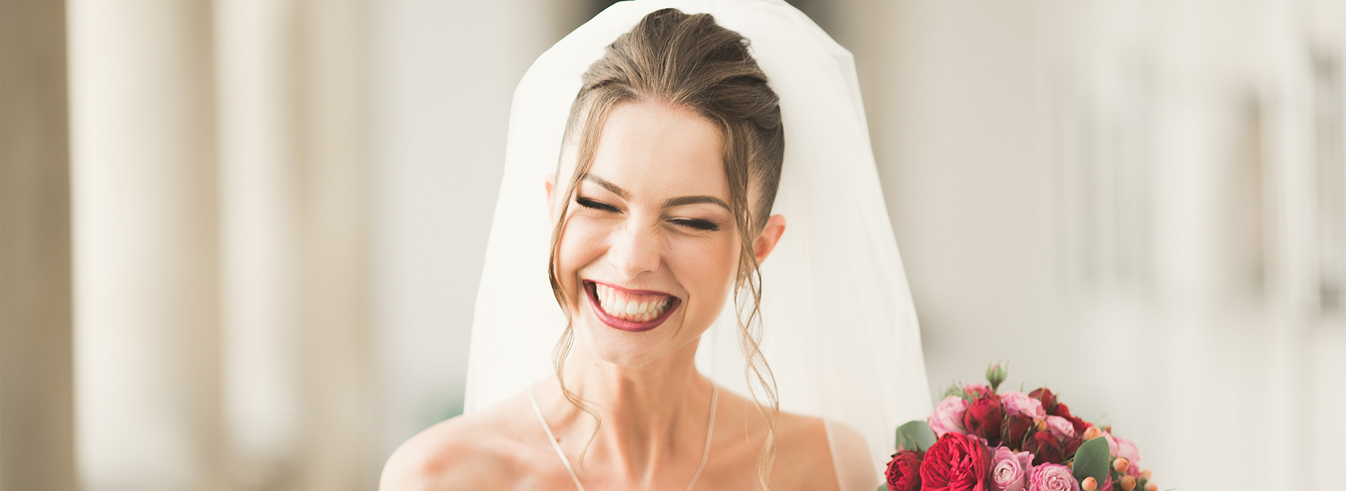 The image shows a woman wearing a wedding veil and smiling, with her hand near her mouth. She is standing in front of a blurred background that suggests an indoor setting, possibly a church or reception hall.