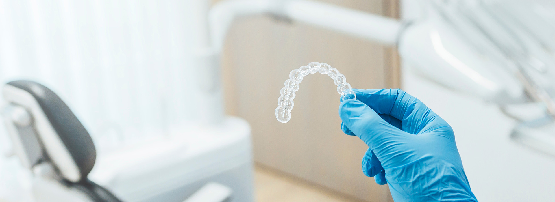 An image of a dental professional wearing gloves and holding a transparent mold, set against a sterile background with medical equipment.