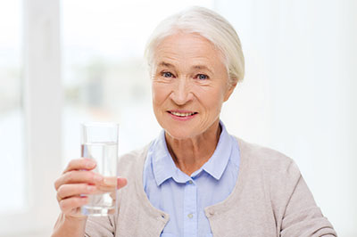 The image shows an elderly woman holding a glass of water, smiling and appearing content.