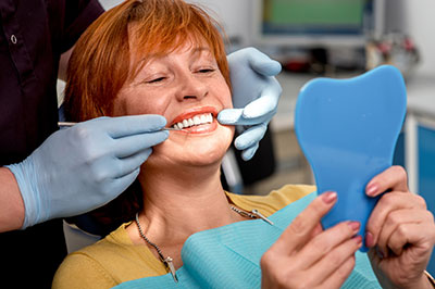 A woman sitting in a dental chair, smiling at the camera while holding a blue mouth guard, with a dental professional applying it to her teeth.