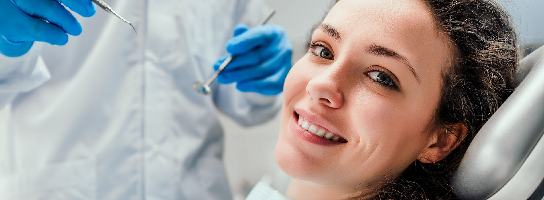 A woman sitting in a dental chair with a smiling expression, receiving dental care from a professional wearing gloves and a mask.