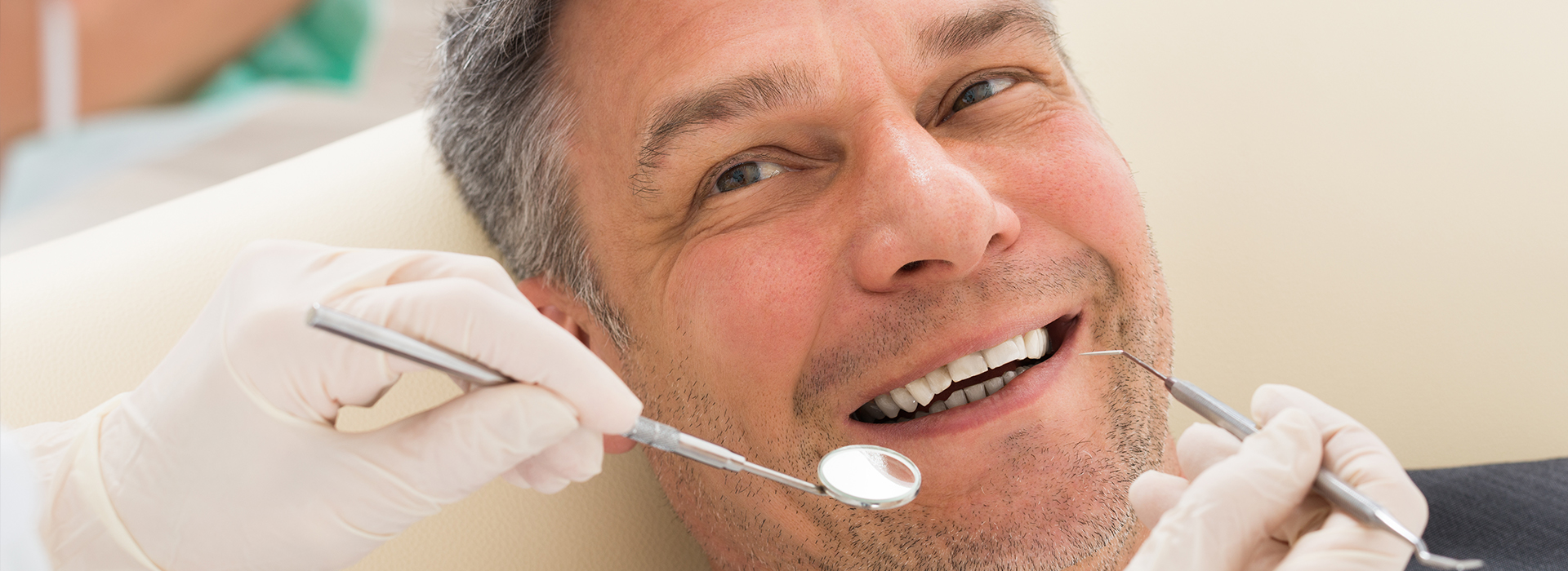 A man is sitting in a dental chair, smiling at the camera, while receiving dental treatment with a dentist working on his mouth.