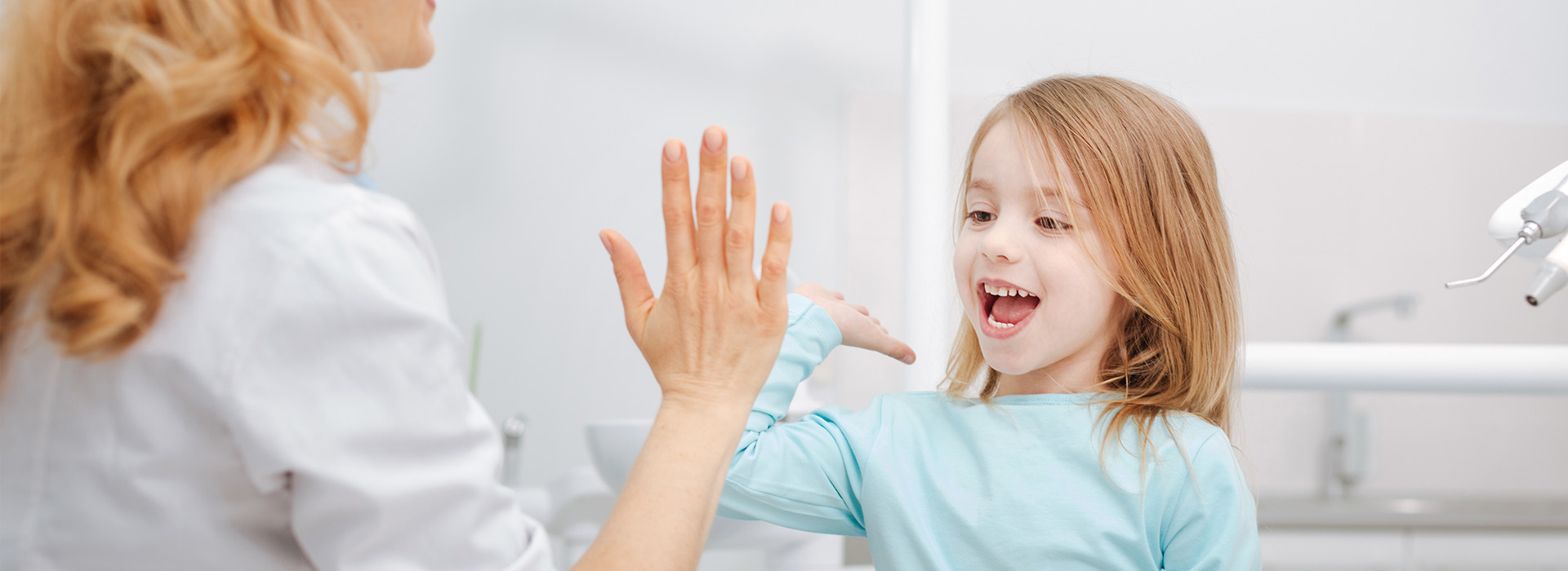 The image is a photograph of two individuals with one person, presumably an adult female, interacting with the other, who appears to be a young child. They are in what seems to be a dental or medical setting, as indicated by the presence of a sink and a chair designed for patient use.