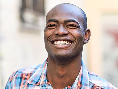The image shows a smiling man with short hair, wearing a plaid shirt and standing outdoors.