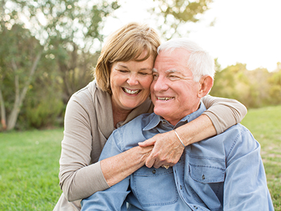 The image shows an elderly couple, a man and woman, embracing each other in a warm pose. They are outdoors during the daytime.