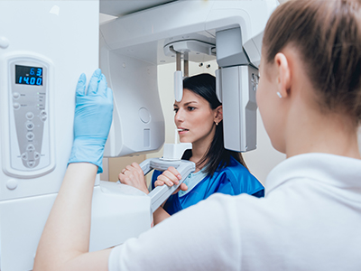 A woman in a white lab coat is standing next to a large, modern scanner machine with a digital display, while another person in a blue shirt and face mask examines the device.