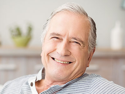 The image shows an older man with a smile, wearing glasses and sitting in a relaxed posture.