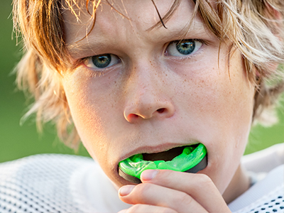 A young boy with blonde hair, wearing a sports jersey and brushing his teeth with a green toothbrush.